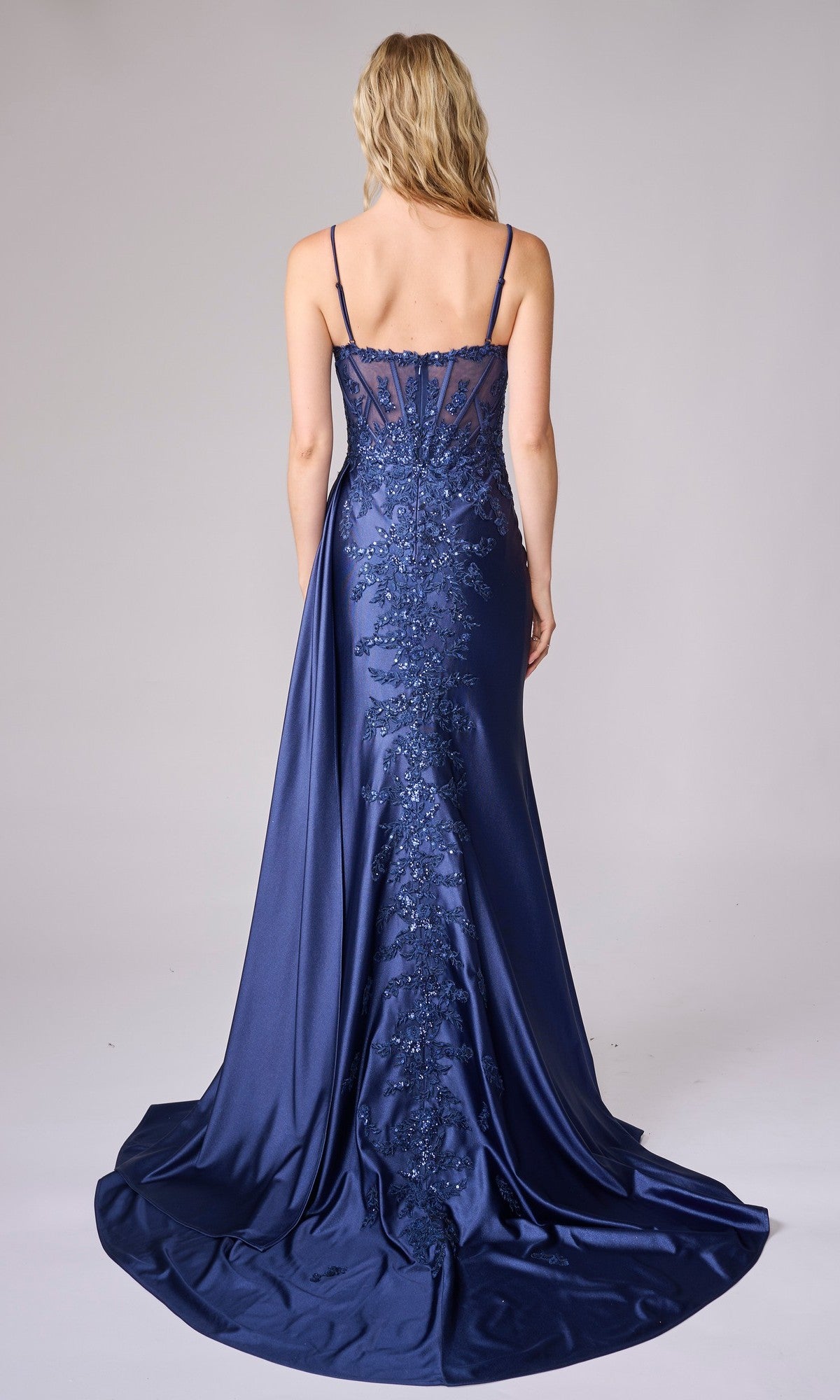 A woman stands with her back to the camera, wearing the Nina Canacci 7655—a floor-length navy blue satin formal dress featuring spaghetti straps and intricate embroidery down the back.